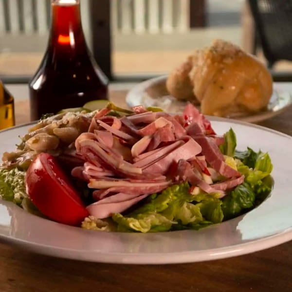 Italian salad with lettuce, tomato, sliced meats, and pasta, with a bottle and bread in the background.